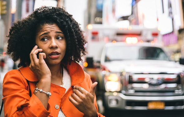 Woman on phone looking concerned, ambulance in background on a city street.