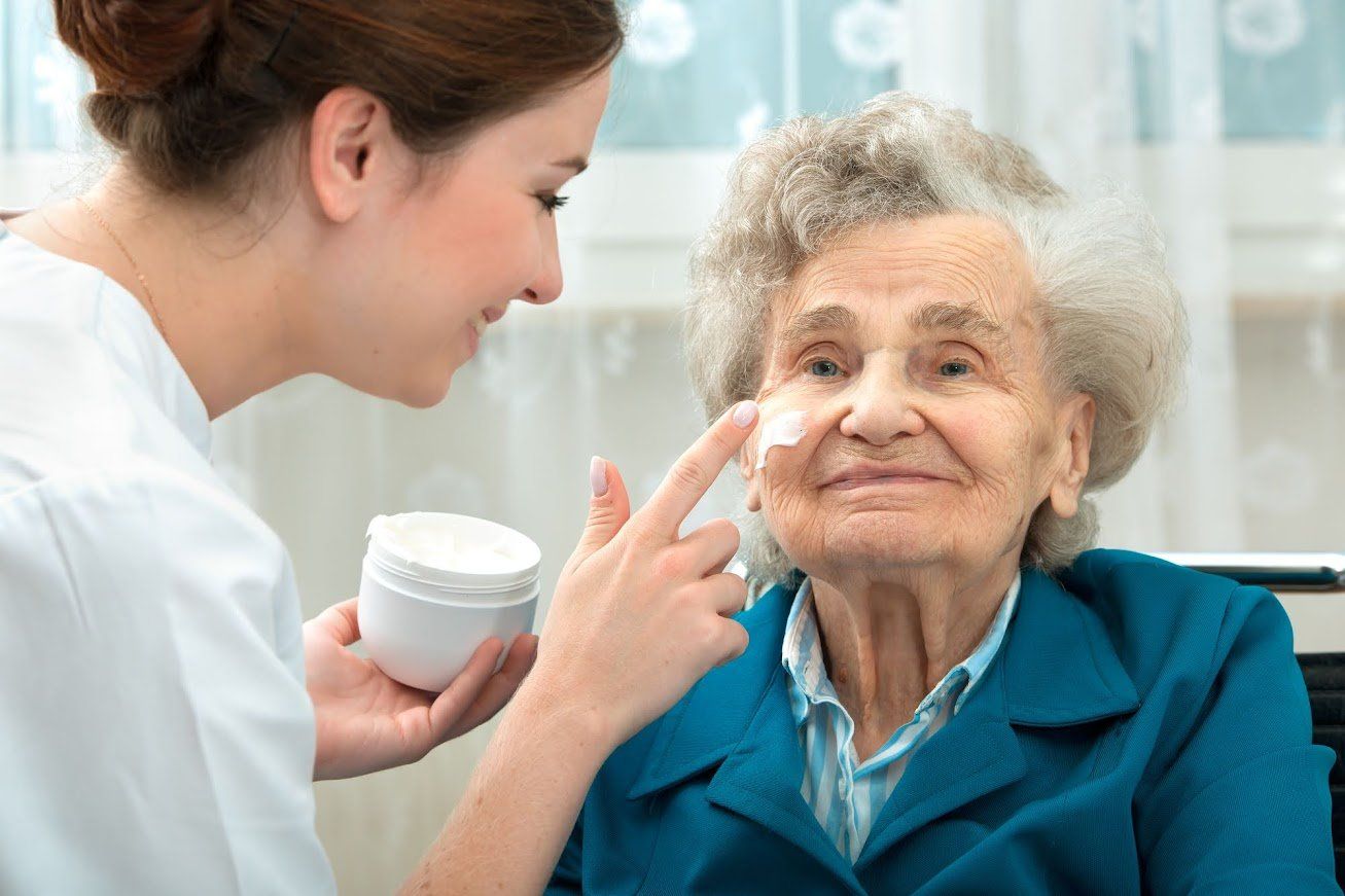 Caregiver applying cream to an elderly woman's face, indoor setting.