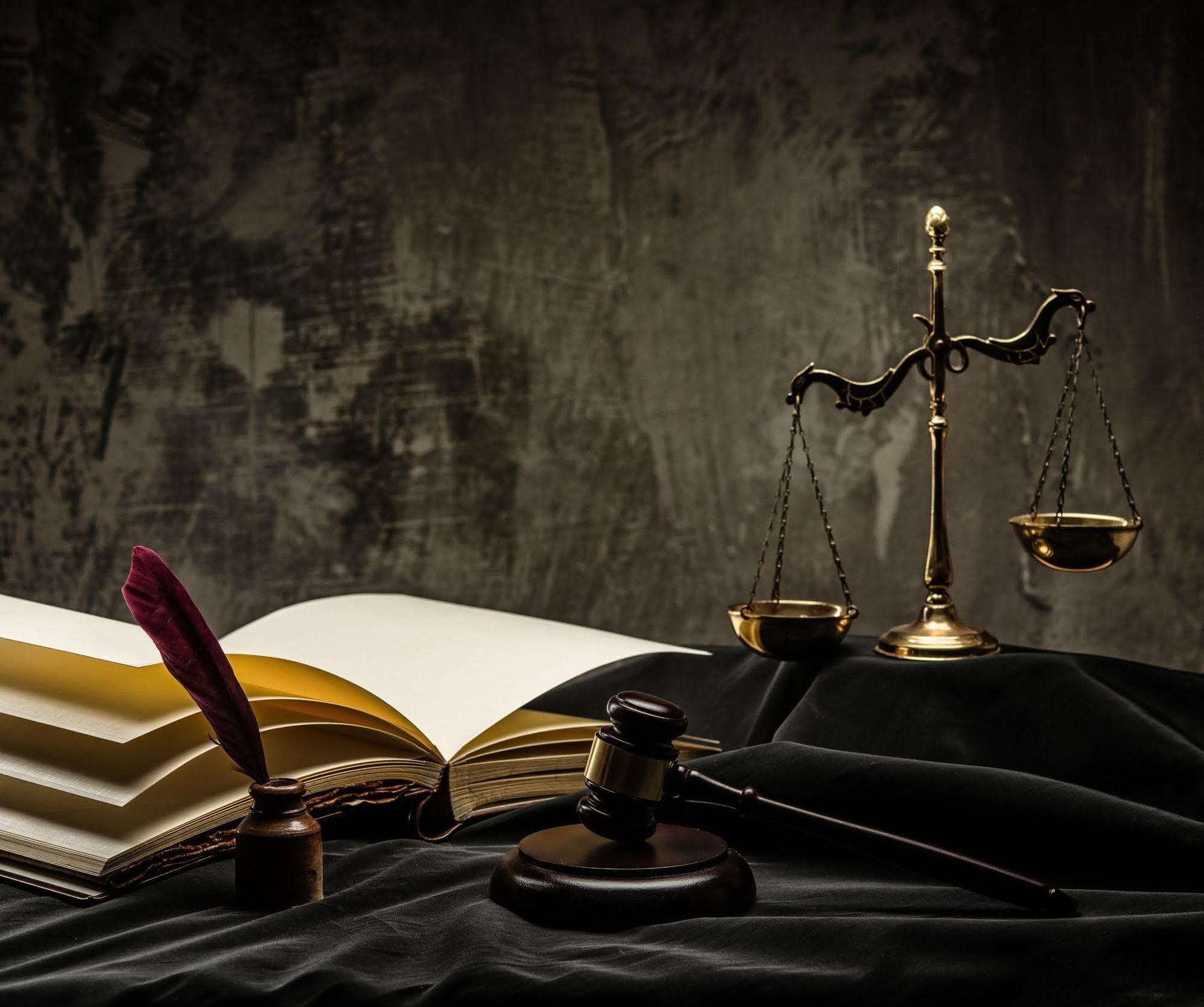 Scales of justice, gavel, open book, inkwell, and quill on a black cloth, dark textured background.