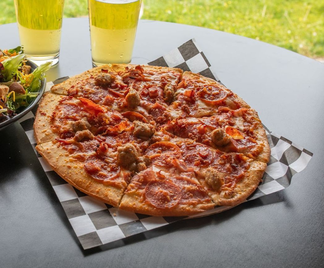 Pizza with sausage and pepperoni on checkered paper, two beers, and a salad on a table.
