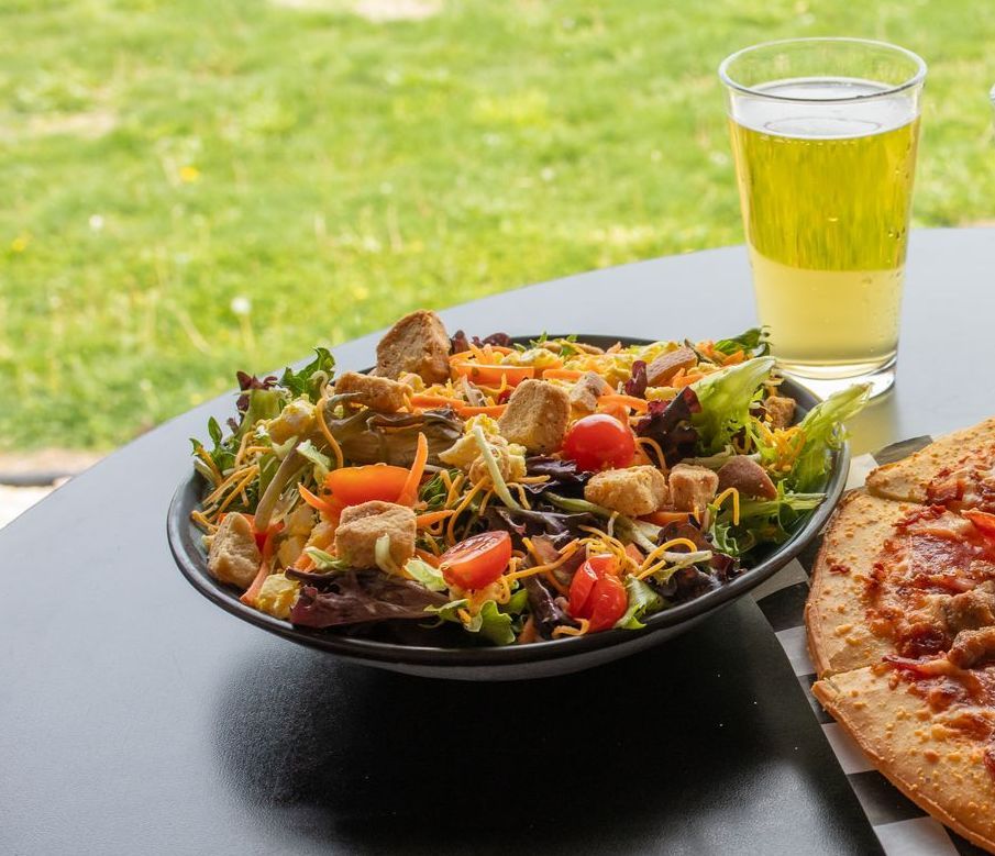 Salad with tomatoes, cheese, and croutons, glass of yellow drink, and part of pizza on black table, outdoors.