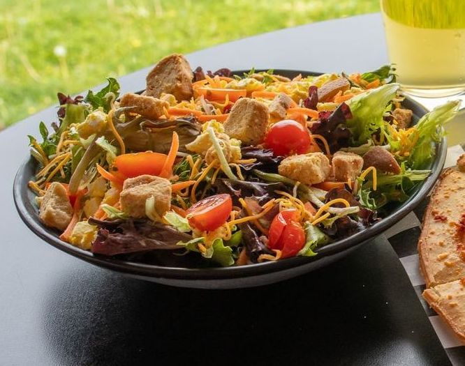 Salad bowl with croutons, tomatoes, shredded cheese, and carrots on a black table.