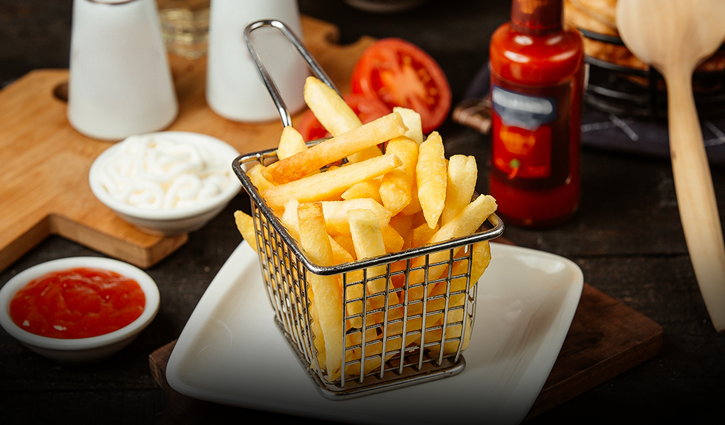 French fries in a wire basket on a white plate, with ketchup, mayonnaise, and tomato slice.