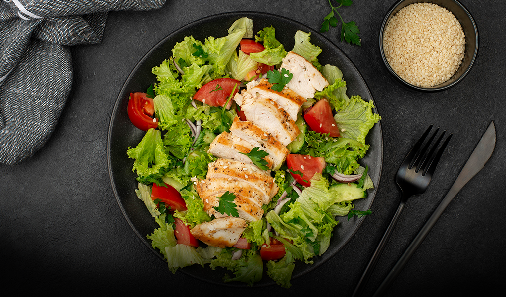 Chicken salad with tomatoes, lettuce, and sesame seeds in a black bowl.
