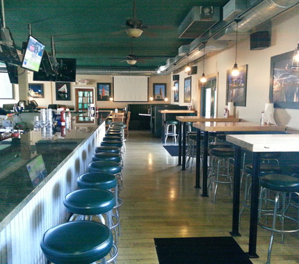 Interior view of a bar with a long counter, stools, tables, and a screen; green and beige color scheme.