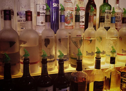 Bottles of liquor lined up on a bar shelf. Many have green pour spouts, showcasing various brands.