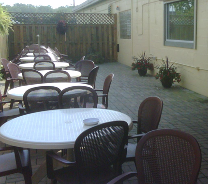 Outdoor dining area with white tables and dark chairs on brick patio. Building wall in background.