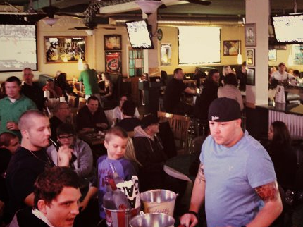 A crowded bar scene; people at tables and bar, TVs on walls, a man in a blue shirt in foreground.