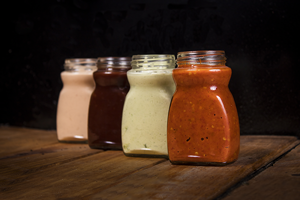 Four jars of sauce in varying colors on a wood surface: beige, brown, green, and red.