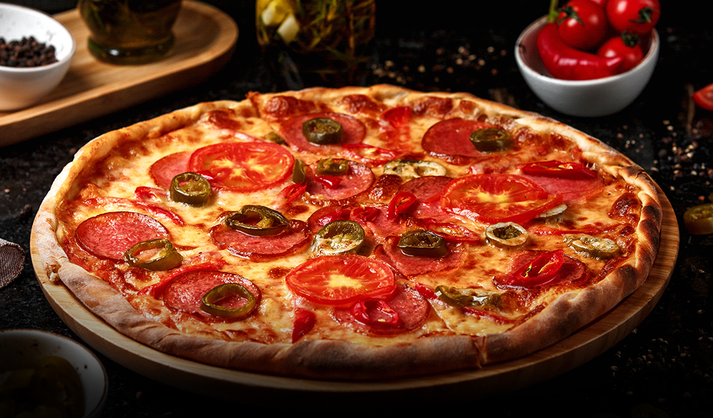 Pizza with pepperoni, tomatoes, and jalapeños on a wooden board. Ingredients visible in background.