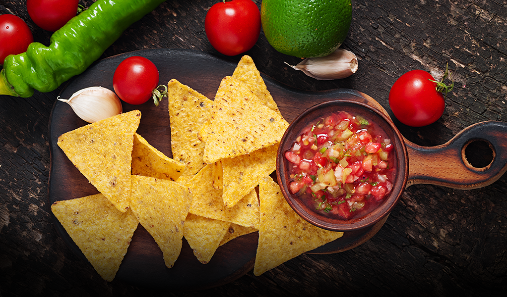 Tortilla chips with salsa in a wooden bowl, surrounded by tomatoes, garlic, peppers, and a lime.
