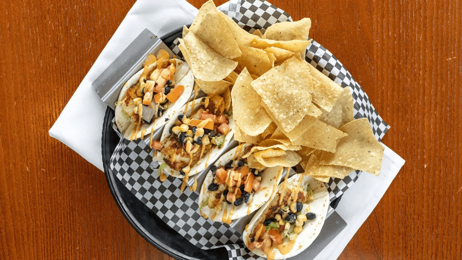 Tacos with black beans, toppings, and chips in a basket on a wood table.