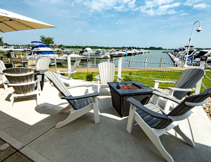 Outdoor patio with Adirondack chairs around a fire pit, overlooking a marina on a sunny day.