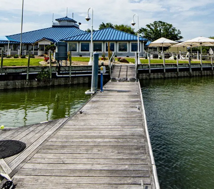 Wooden dock leads to a waterfront building with blue roof. Umbrellas and grass surround.