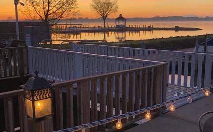 Wooden deck with a lit lantern, overlooking a calm lake at sunset with a gazebo.