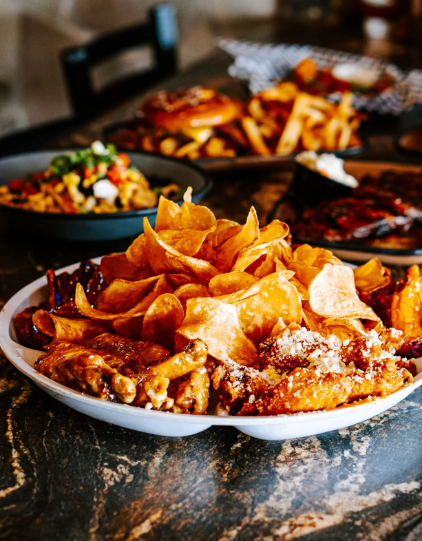 Close-up of a plate of potato chips and chicken wings; other dishes and a burger are visible in the background.