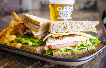 Sandwich with fries, lettuce, tomato, bacon, and cheese on a plate, with a beer in the background.