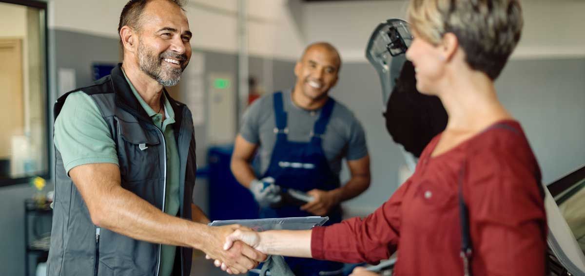 A woman shaking hands with a mechanic in an auto shop, another mechanic smiles in the background.