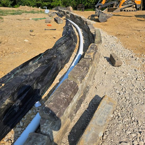 Construction site: white pipe in trench, surrounded by rocks and gravel. Backhoe in background.