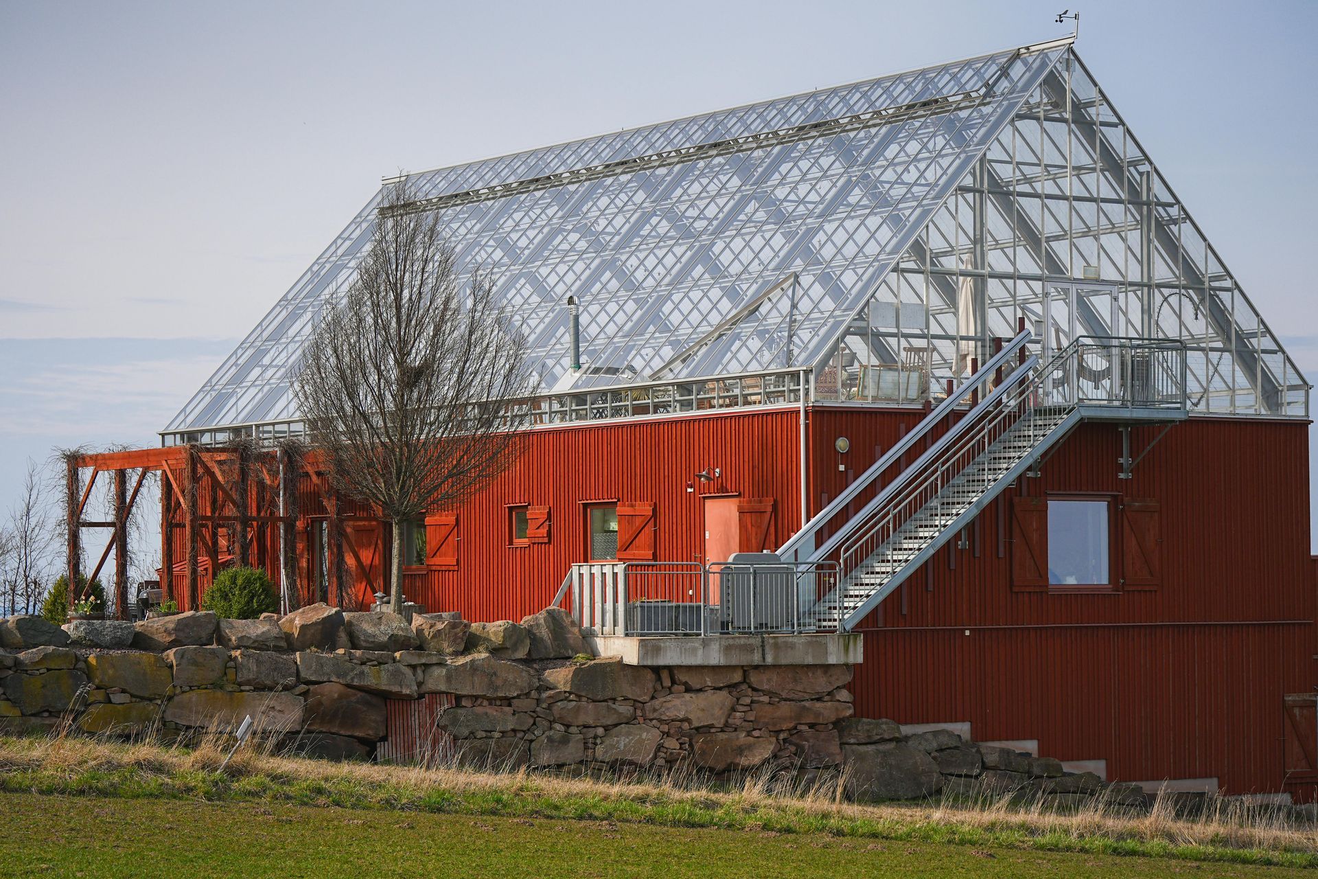 Red barn with a large glass greenhouse roof on a stone foundation in a grassy field.