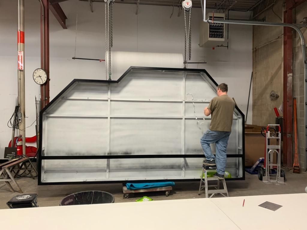 Man on step stool working on a large white rectangular frame with black trim; industrial setting. Man on step stool working on a large white rectangular frame with black trim; industrial setting.