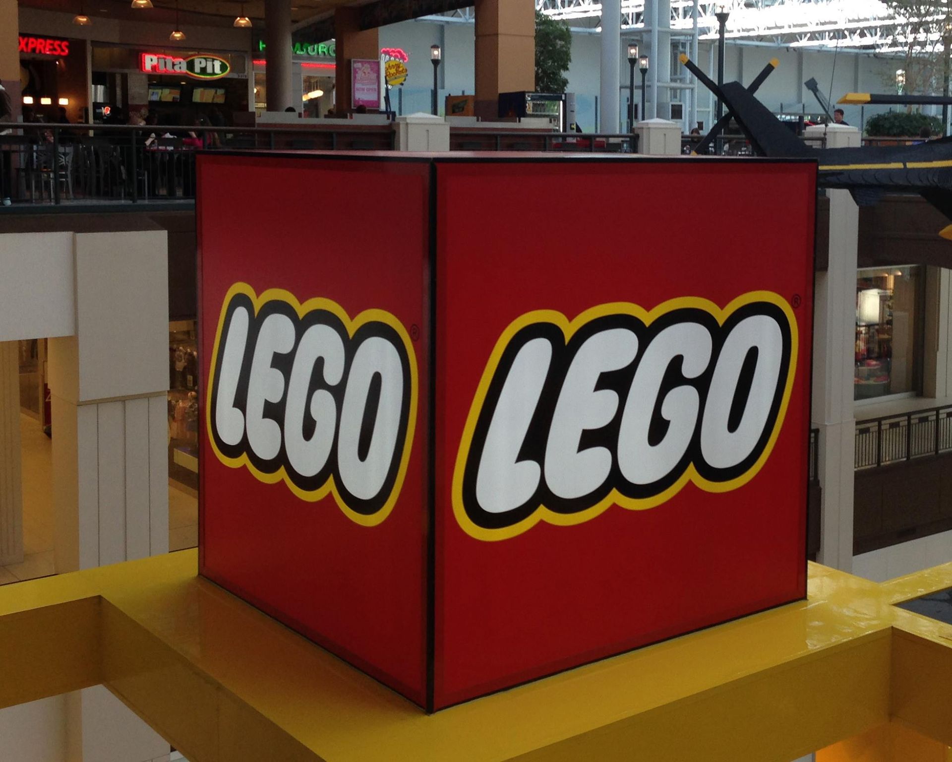 Large red LEGO cube with white text and yellow border, in a mall setting. Large red LEGO cube with white text and yellow border, in a mall setting.
