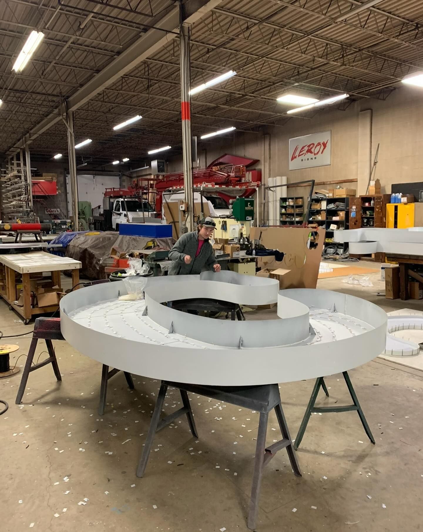 Man in workshop near a large, circular, light-colored sign under construction. Man in workshop near a large, circular, light-colored sign under construction.