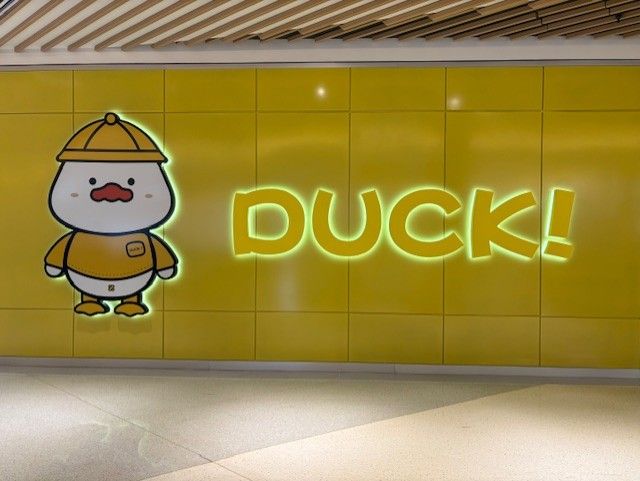 Cartoon duck mascot beside glowing yellow “DUCK!” sign on a tiled wall