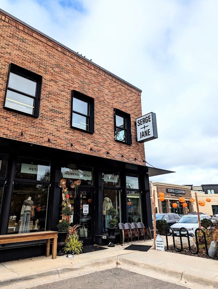 Brick storefront with black-trimmed windows, a hanging sign, and an outdoor café table on a sidewalk corner.
