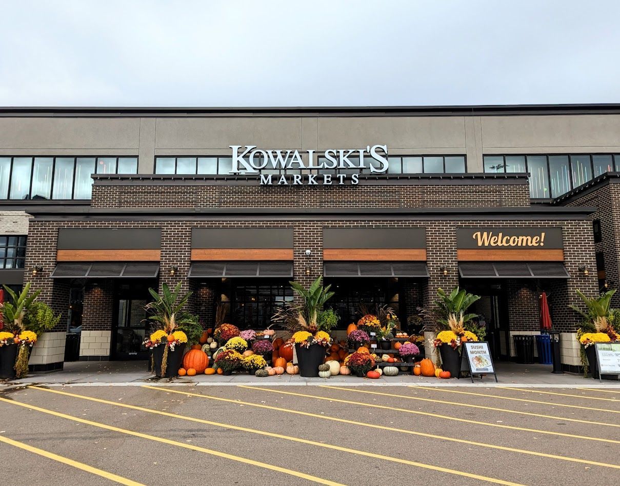 Storefront decorated with pumpkins and fall flowers, with a “Welcome” sign and yellow-striped parking lot.