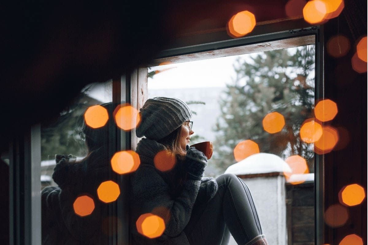 Person in a knit hat sits by a window, looking out at snowy scenery with warm orange lights in the foreground