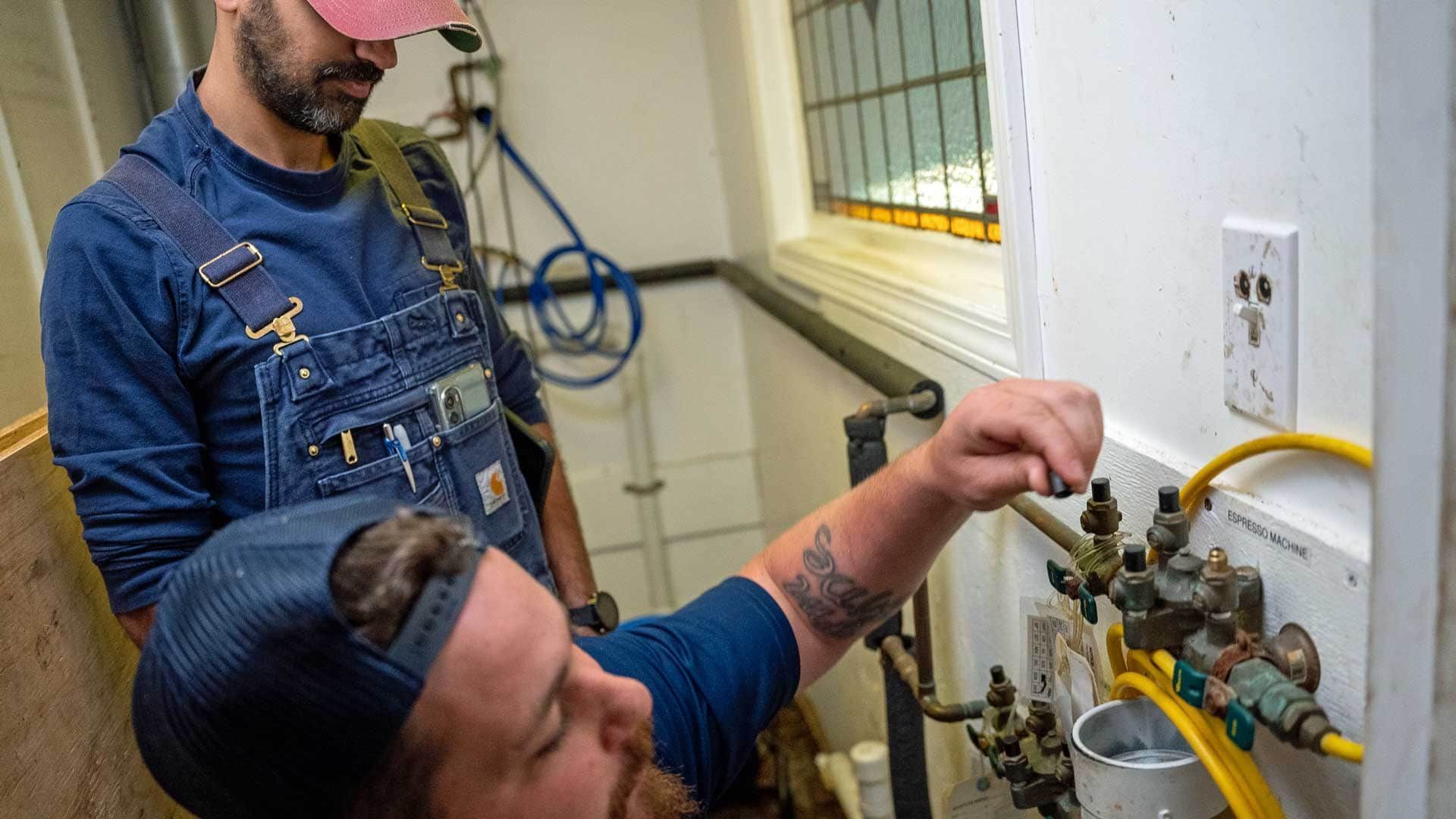Two workers inspecting a wall-mounted gas meter and valves in a utility room