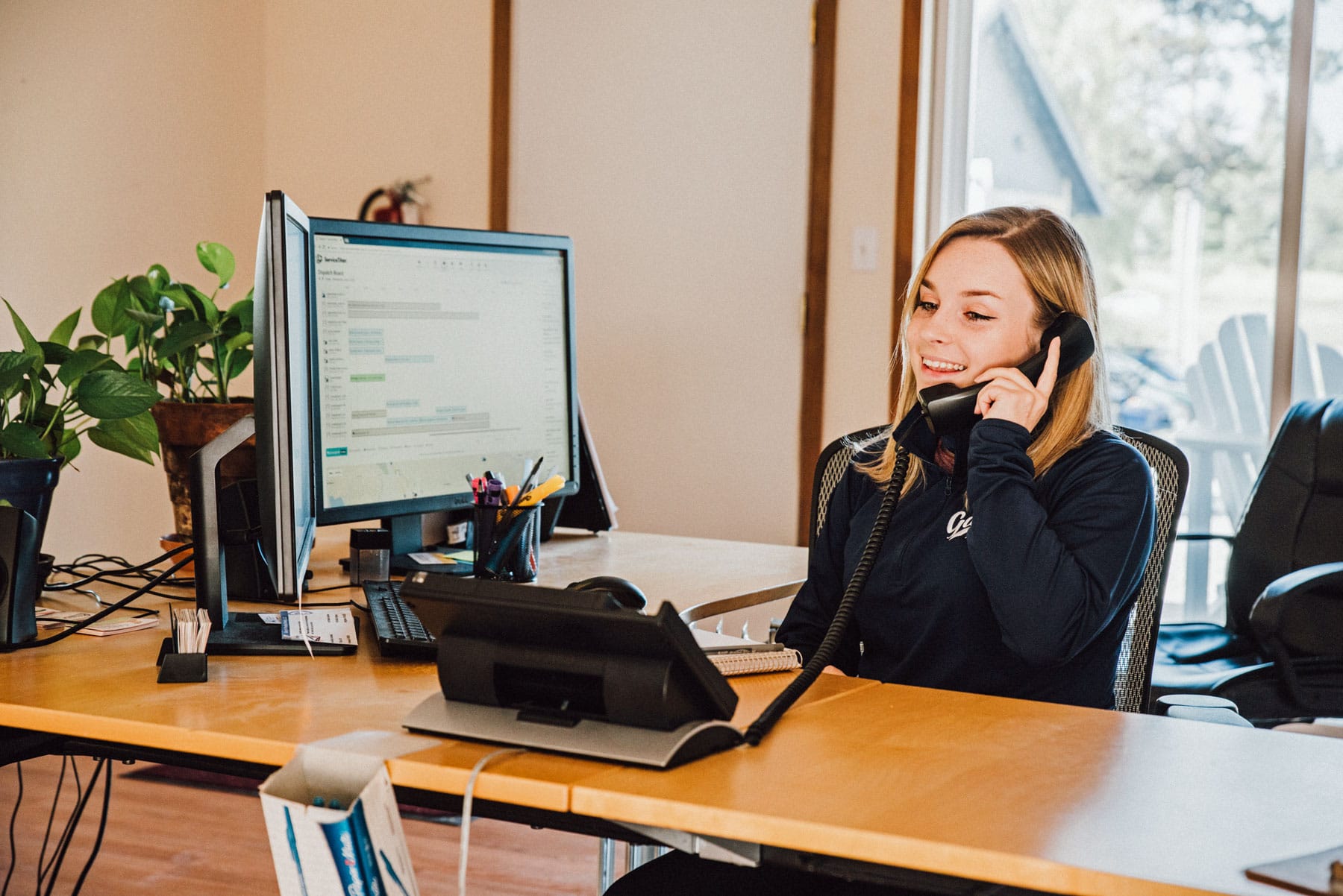 Person on the phone at a desk in an office, smiling beside a computer and laptop.
