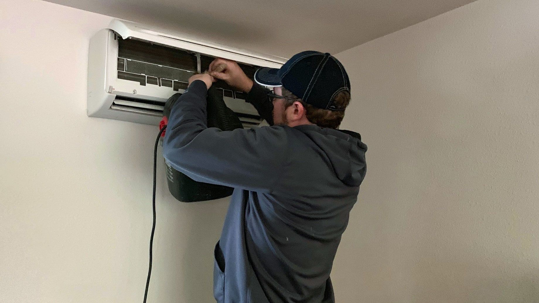 Person in cap using a drill to work on a wall-mounted air conditioning unit indoors.
