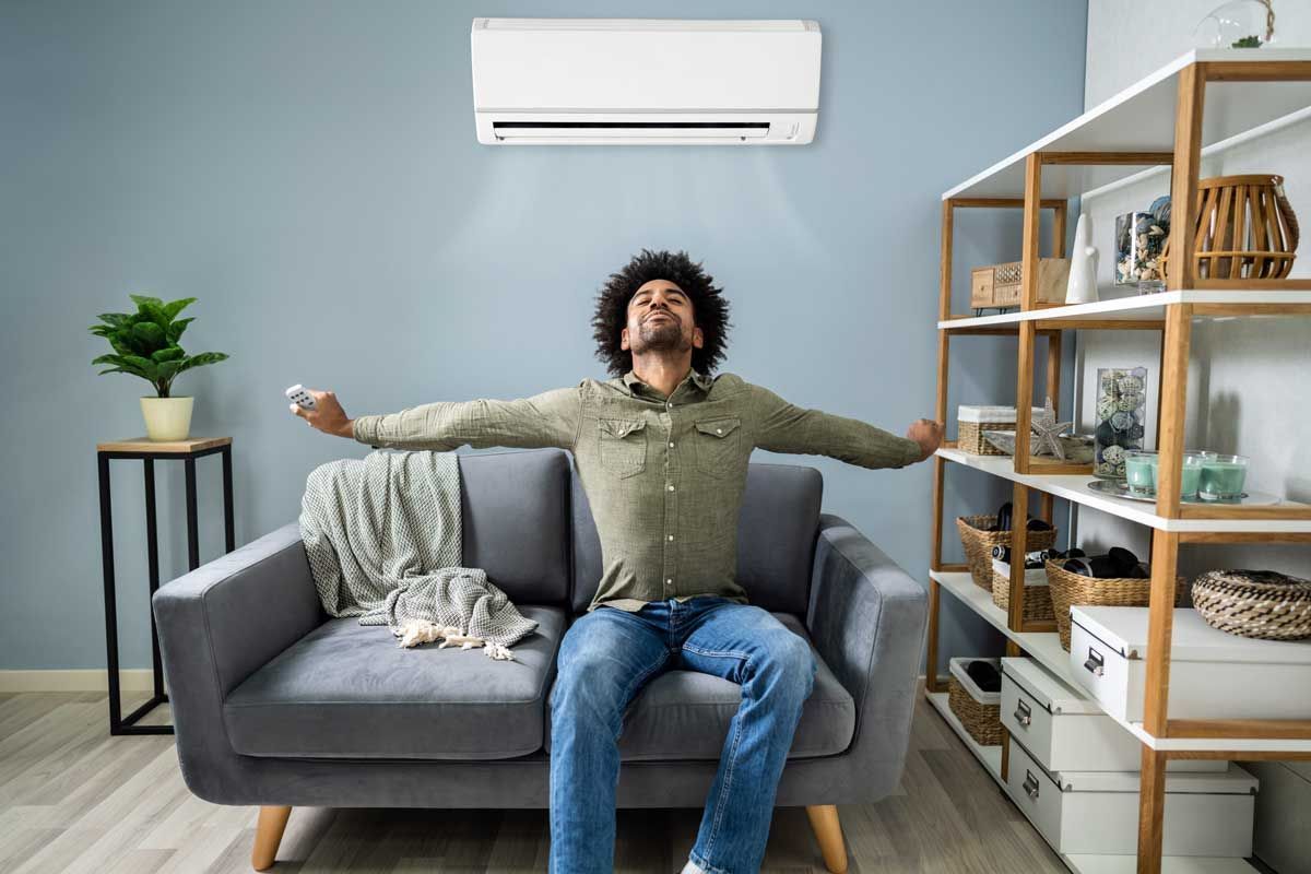 Person lounging on a gray sofa with arms outstretched in a bright living room with an air conditioner above.