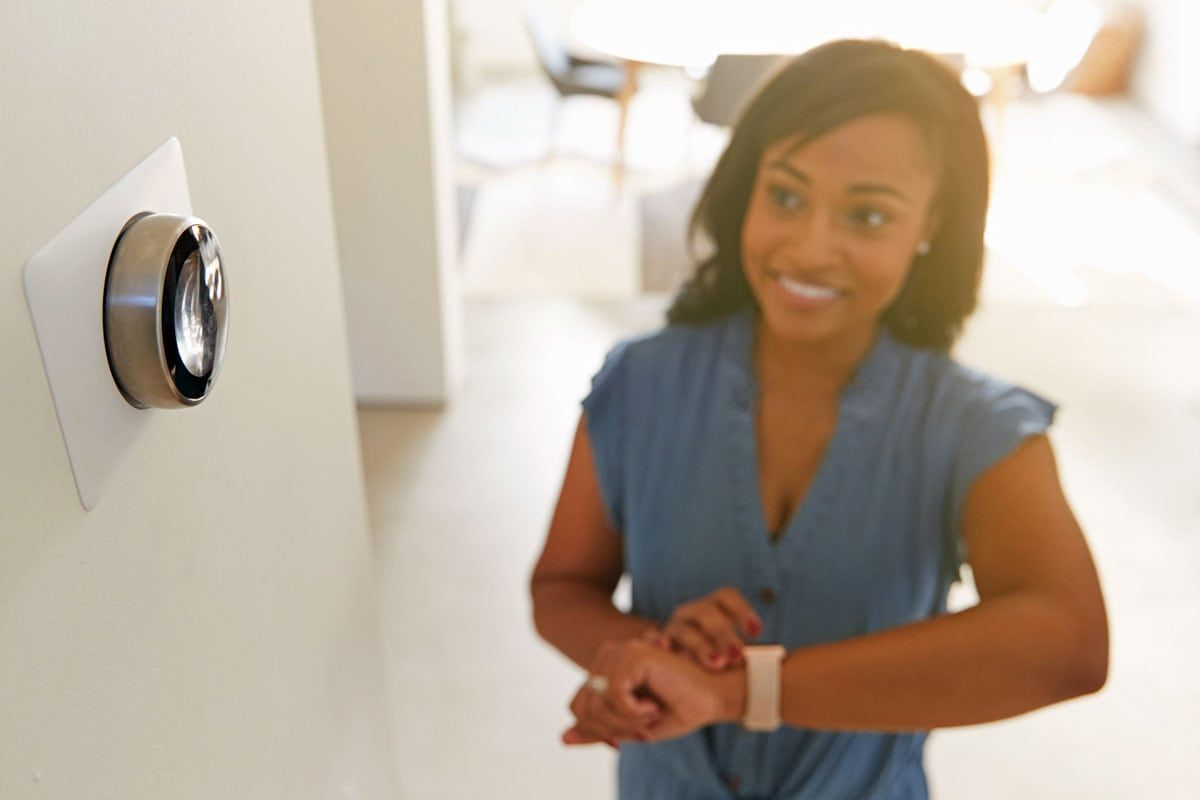 Woman smiling at a wall-mounted thermostat in a bright hallway