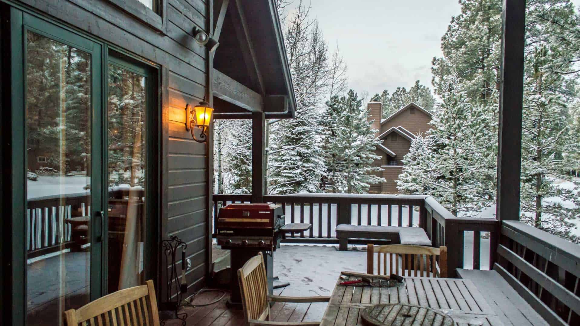 Snowy cabin deck with wooden chairs, railing, and warm porch light overlooking a winter view