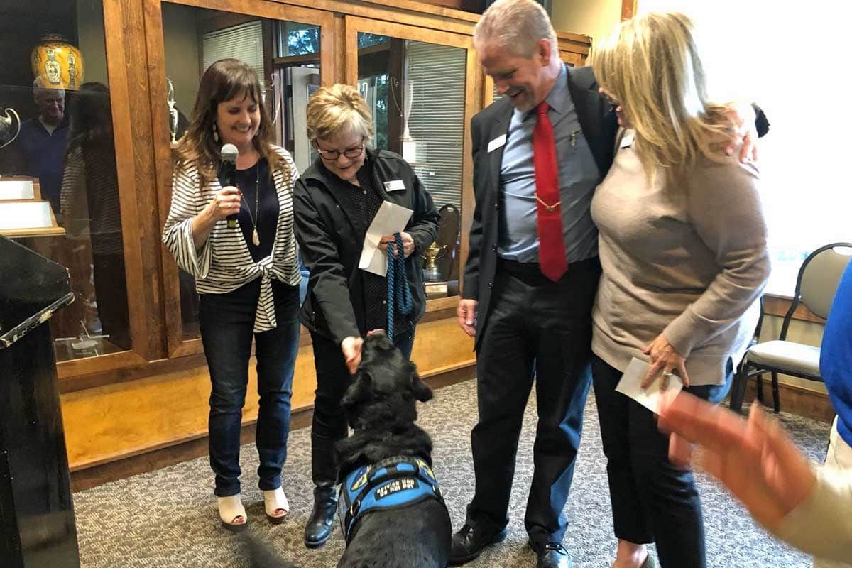 Four people indoors greet a black dog in a blue harness near a doorway.