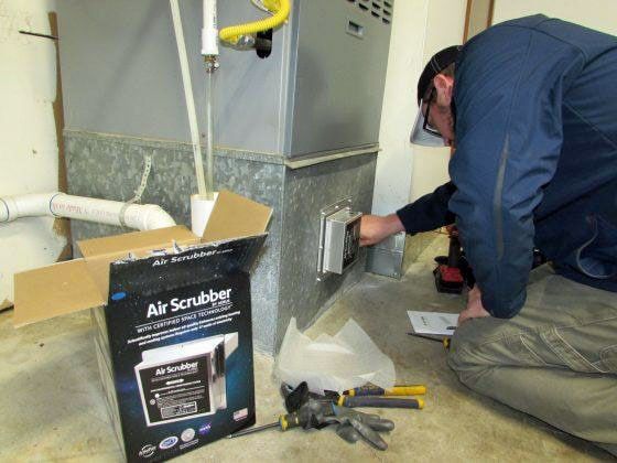 Worker kneels beside furnace, installing an air scrubber in a basement utility room.