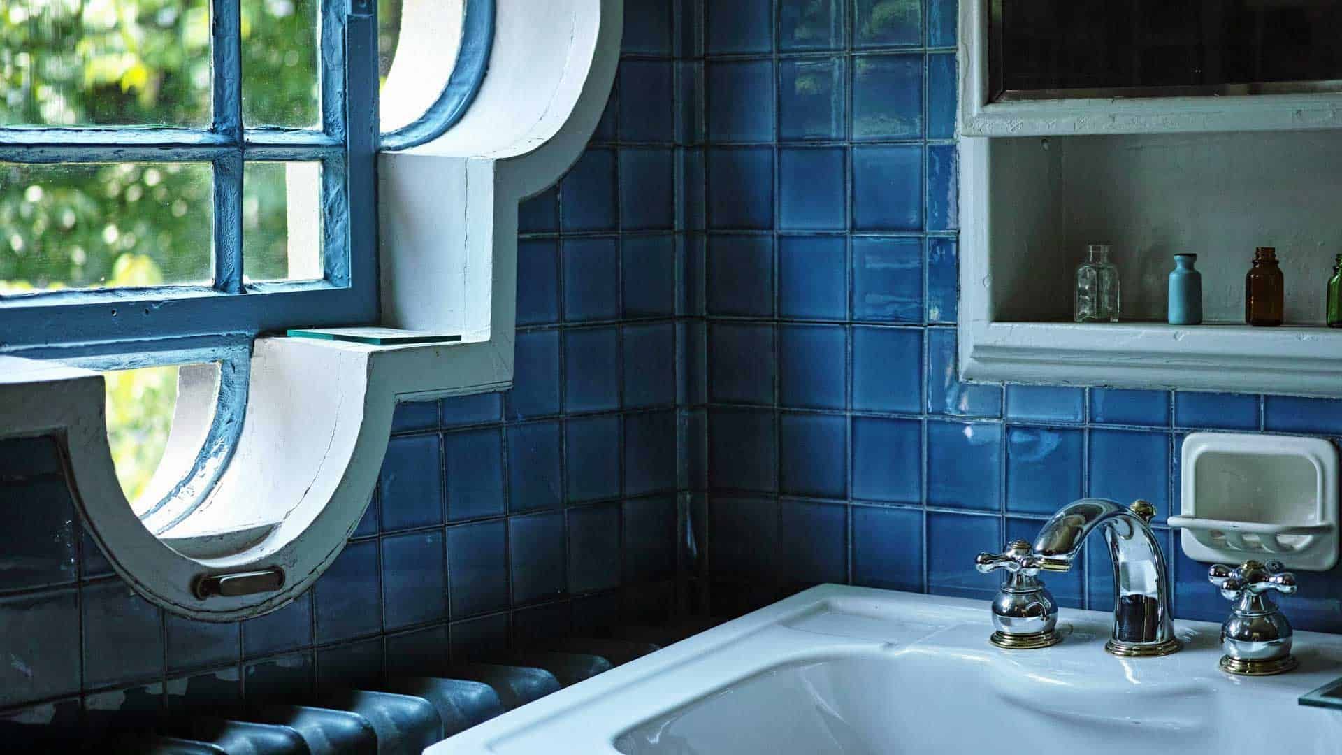 Blue tiled bathroom with a sink, round window, and wall shelf of toiletries