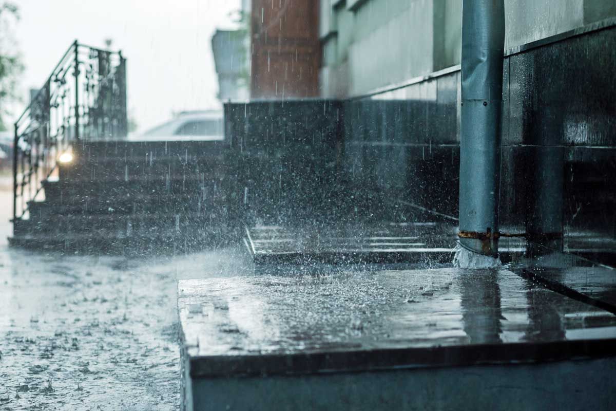 Rain pouring onto wet steps and pavement beside a building, with water splashing on a ledge