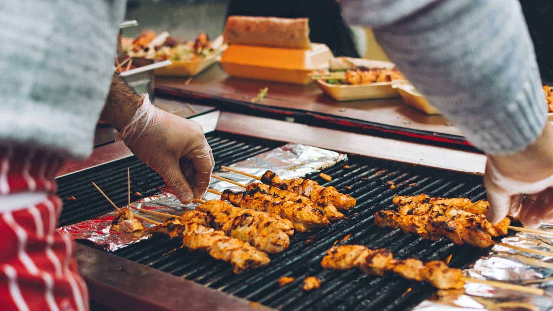Hands grilling skewered meat over a charcoal barbecue outdoors.