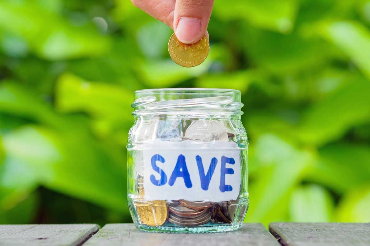 Hand placing coin into a glass jar labeled “SAVE” on a wooden surface with green background