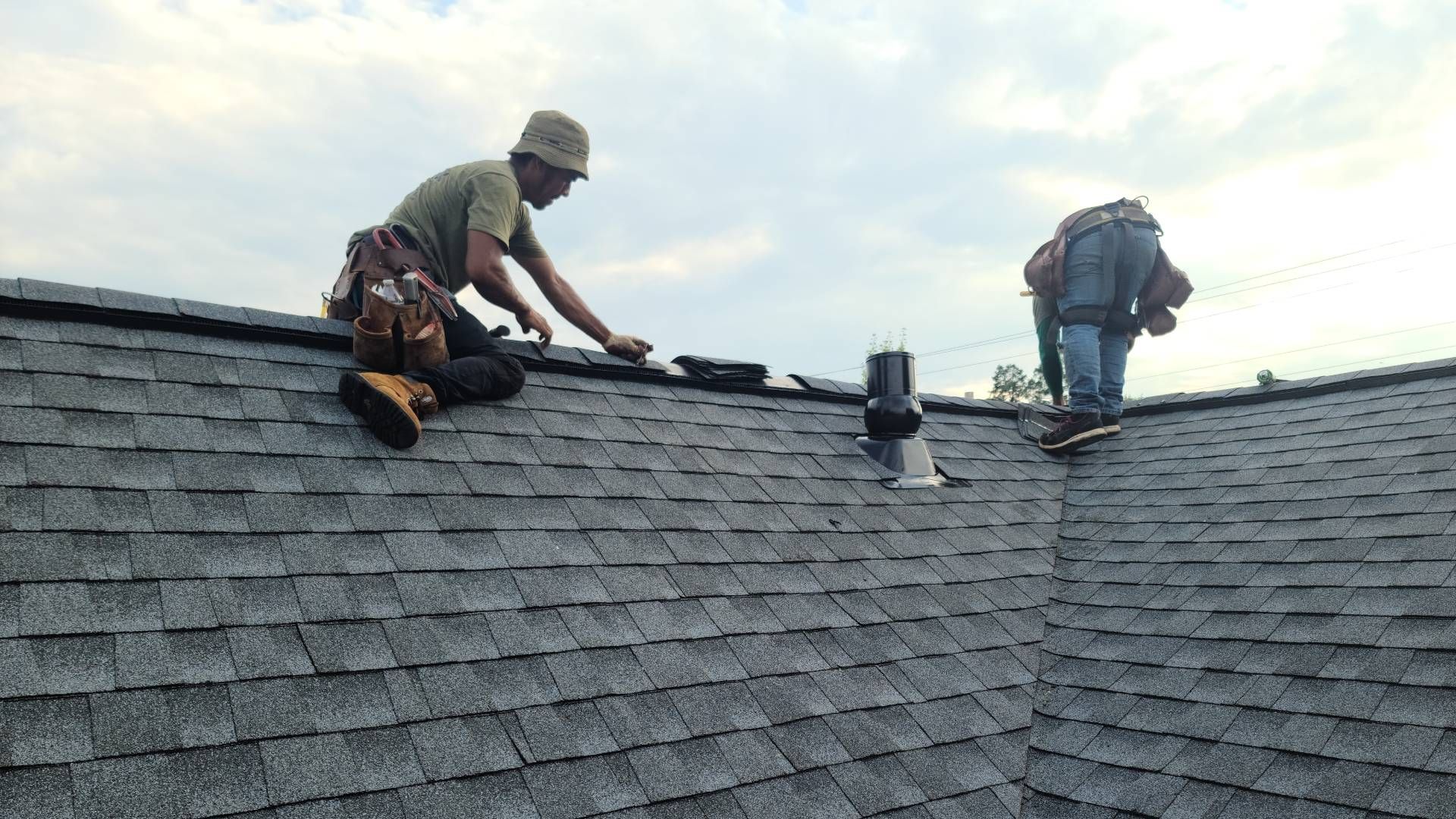 Two roofers installing shingles on a gray shingled roof under a cloudy sky.