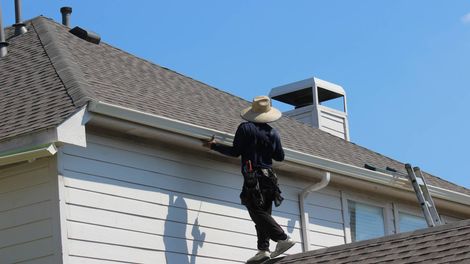 Person on roof cleaning gutters on a house. Blue sky, white siding, brown roof.