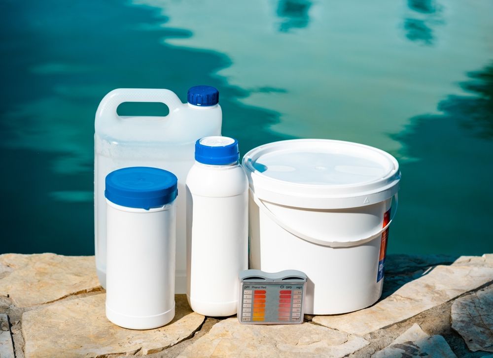 White plastic chemical containers with blue caps and a bucket beside a pool