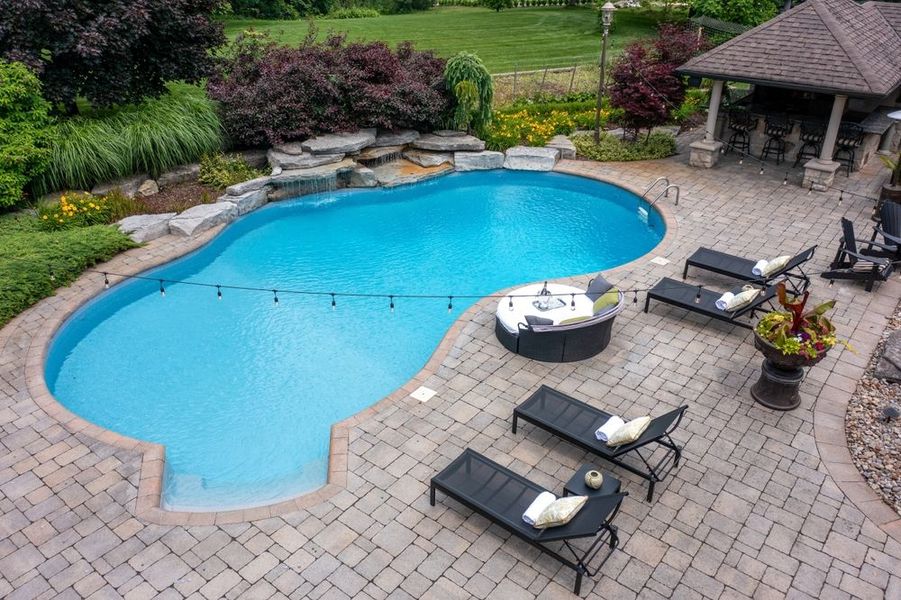 Backyard pool with stone patio, lounge chairs, and hot tub surrounded by greenery