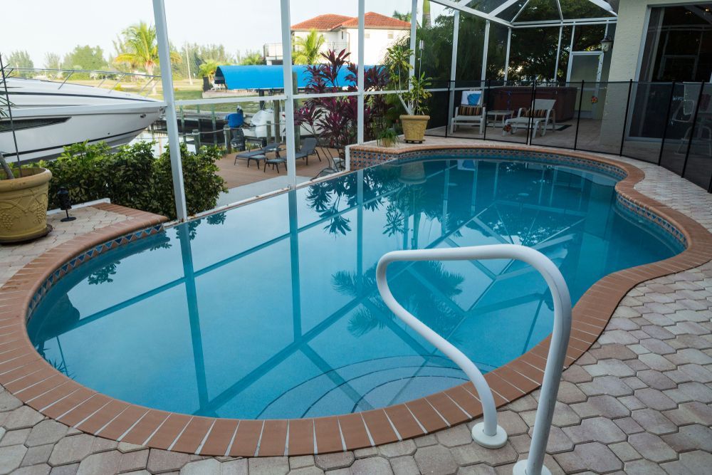 Backyard pool with brick deck and covered patio, featuring a metal handrail and calm blue water