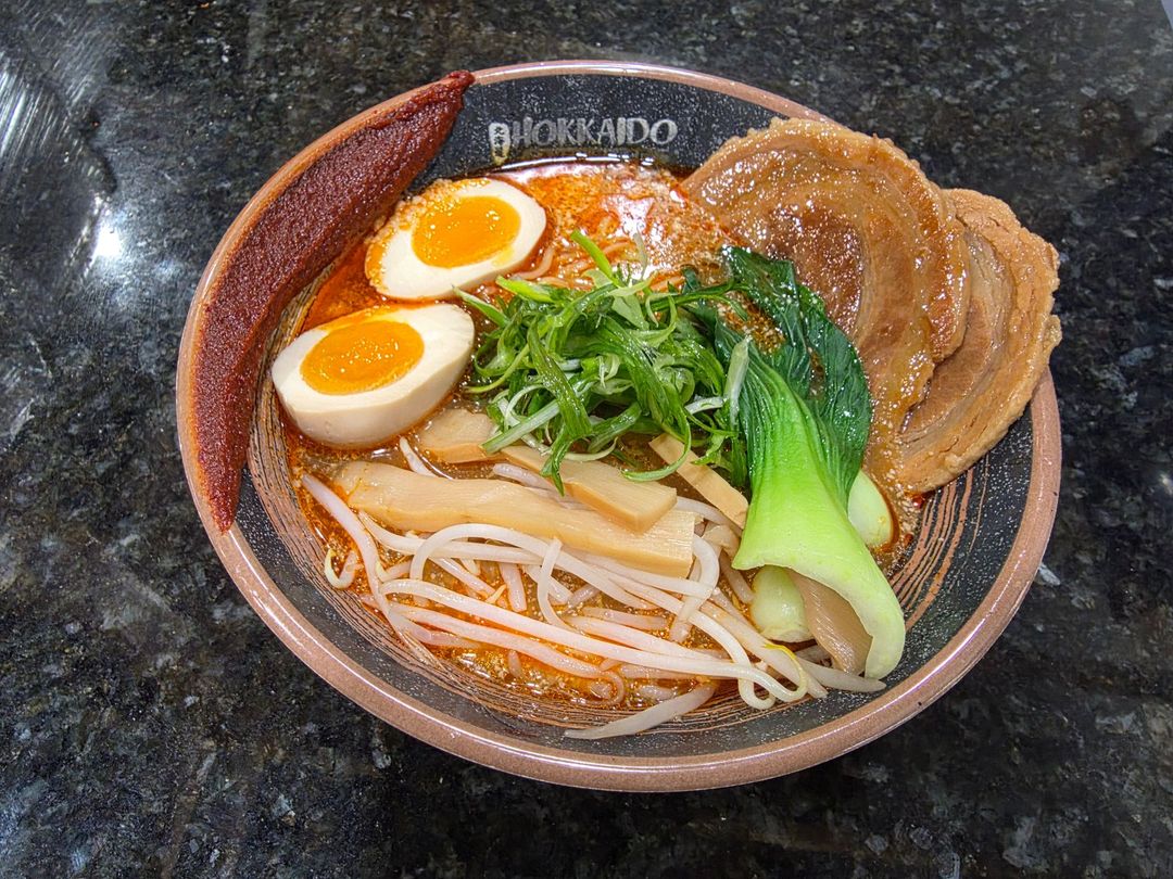 A bowl of ramen topped with mussels, shrimp, soft-boiled eggs, nori, narutomaki, and bamboo shoots on a dark table.