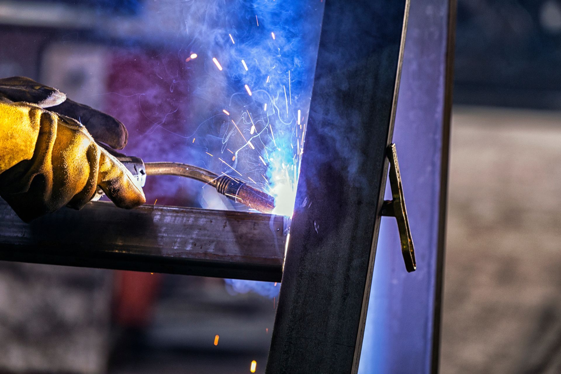Gloved hand welding a metal frame, with bright blue sparks in a workshop